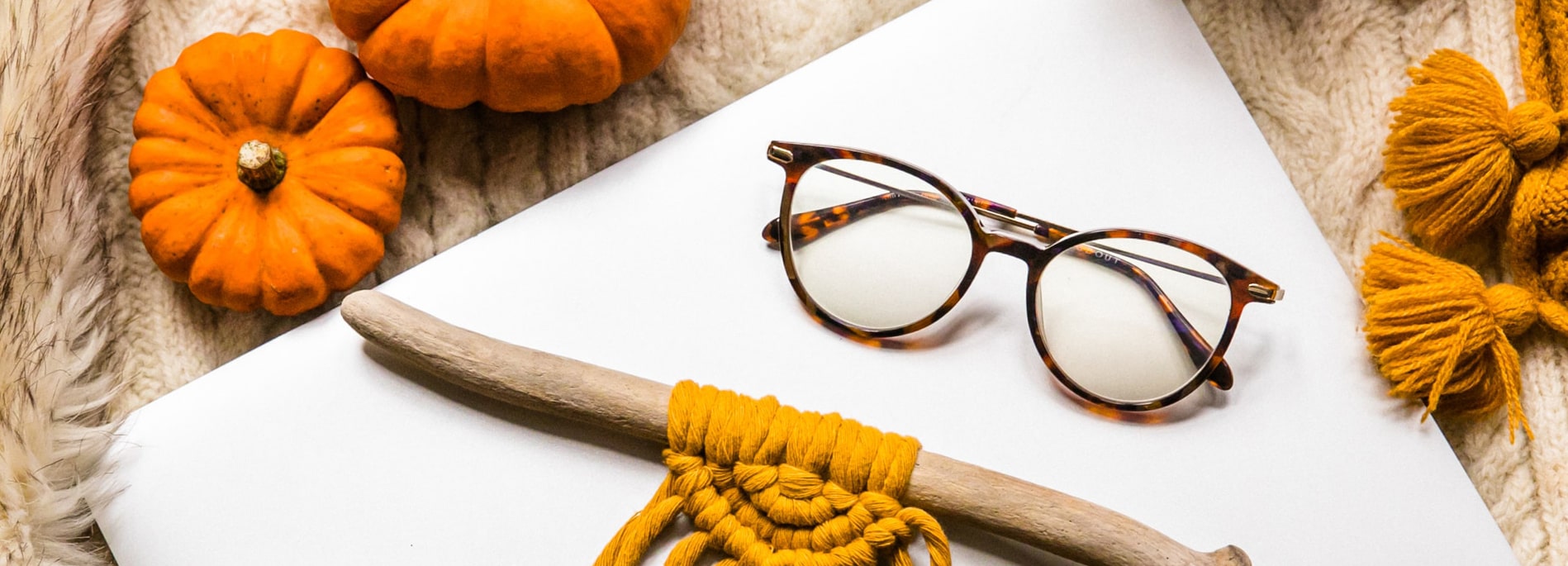 A pair of round tortoiseshell glasses rests folded on white paper, surrounded by pumpkins and cosy beige and yellow knits.