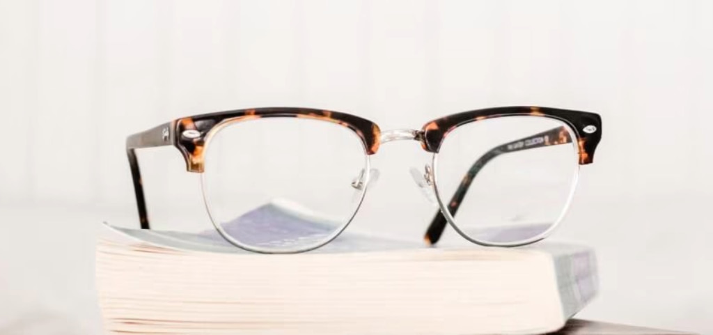 A pair of Clubmaster glasses with a mix of Havana acetate and gold metal positioned at an angle on  top of a book. The backdrop is blurred white. 