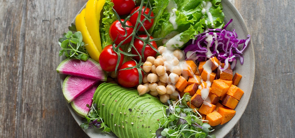 A bowl of colourful, healthy fruit and vegetables, including chopped sweet potatoes, avocado, chickpeas, tomatoes, yellow peppers and lettuce.  