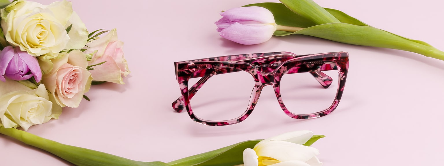 Pink floral patterned square women’s glasses on a soft pink background surrounded by roses and tulips, styled for a Mother’s Day gift guide.