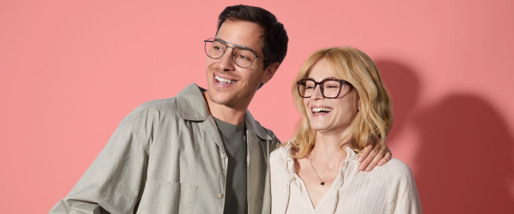 Two models wearing glasses, one male with short dark hair wearing metal aviators and one female with medium blonde hair wearing square tortoiseshell frames, smiling side by side, against a light pink backdrop.