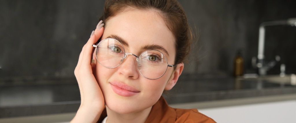 A young woman with hazel eyes gazes directly into the camera, her brown hair gathered into a messy bun. Resting her head gently on one hand, she wears round silver glasses and a soft orange blouse. A faint, knowing smile touches her lips, giving the portrait a calm and approachable warmth.