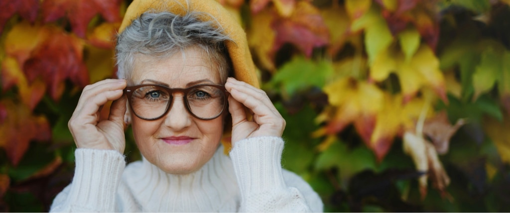 Smiling older woman with short grey hair adjusts round brown acetate glasses, wearing a mustard-yellow hat and white turtleneck sweater, standing in front of colourful autumn leaves.