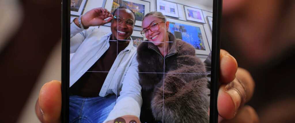 Smartphone screen showing two friends in glasses smiling for a selfie, with camera gridlines visible and hands holding the phone in the foreground.
