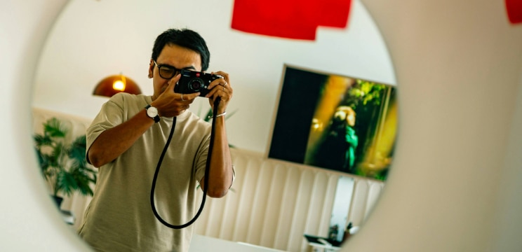 Man with short dark hair wearing angular glasses taking a mirror selfie with a camera, reflected in a round mirror with a softly lit indoor background.