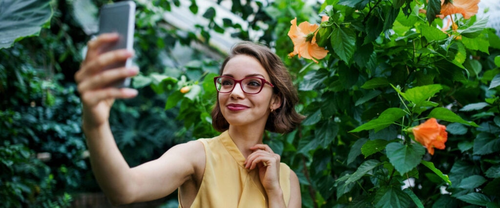 Woman wearing red glasses and yellow sleeveless top, taking a selfie in a lush garden, surrounded by green foliage and orange flowers
