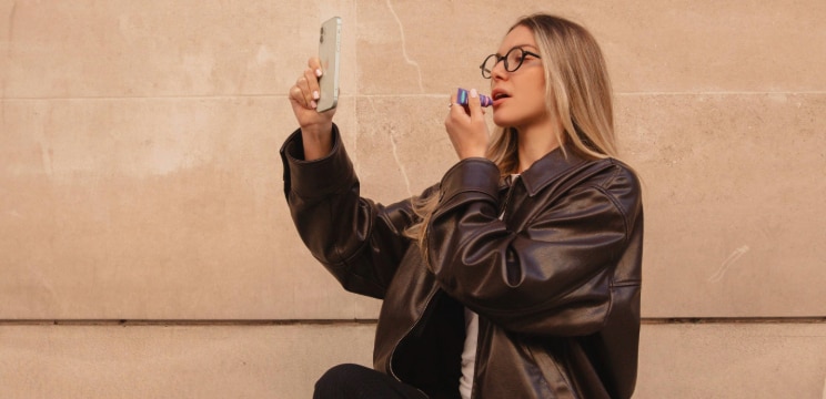 Woman with long straight blonde hair wearing round tortoiseshell glasses applying lip balm while taking a selfie, dressed in a brown leather jacket against a neutral stone wall.