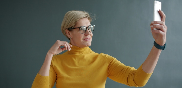 Woman with short blonde hair wearing a yellow turtleneck, wristwatch and dark rectangular glasses taking a selfie on her smartphone against a plain grey background.