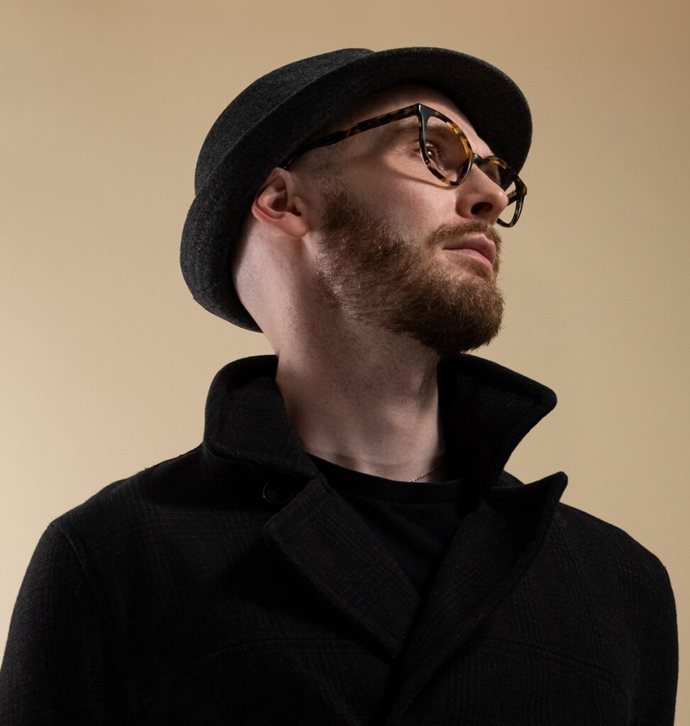 Man posing with a trilby hat wearing tortoiseshell glasses looking to the right