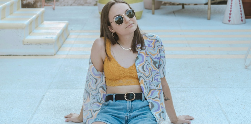 Young woman with medium brown hair sitting on a light-coloured patio in the sun, wearing round gold metal 90s-style sunglasses, a yellow lace bralette, an open pastel patterned shirt, and high-waisted denim shorts.