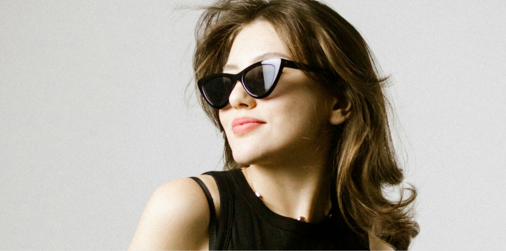 Woman with shoulder-length brown wavy hair wearing black 90s-style black cat-eye sunglasses and a sleeveless black top, looking to the side against a plain light background.