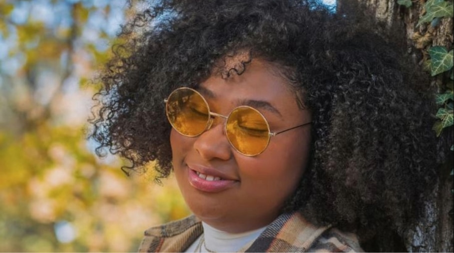 Woman with thick, dark curly hair wearing a plaid shirt and round gold-framed 90s-style sunglasses with amber-tinted lenses, standing beside a tree outdoors with spring foliage in the background.