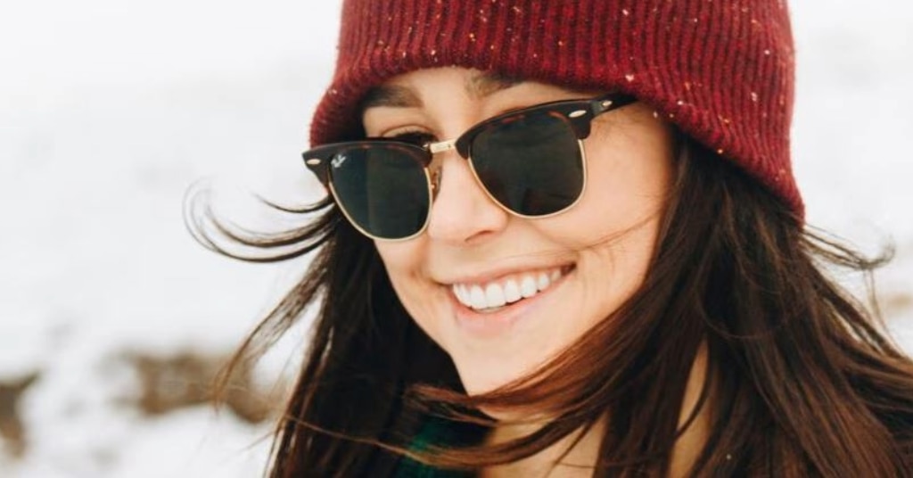 A close up of a young lady wearing a red beanie and Clubmaster sunglasses in a bright winter setting. Her brown hair is blowing slightly in the wind and she is smiling brightly. 