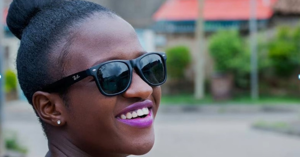 Close-up of a young woman with black hair in a bun, wearing black Ray-Ban Wayfarers in a bright outdoor setting. She smiles brightly with pink lipstick.