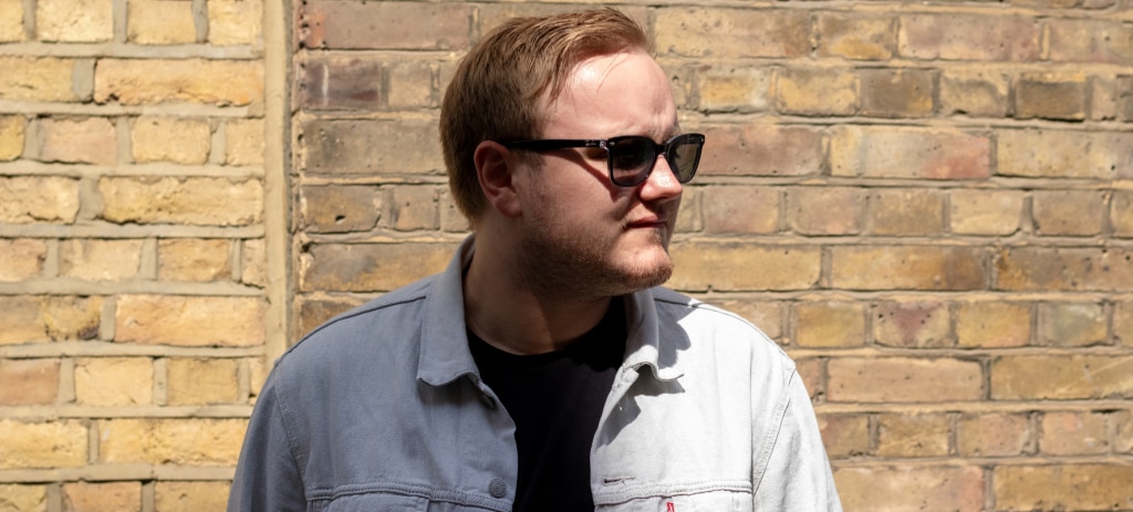 A young man with short blonde hair looks off to his left while standing in front of a yellow brick wall. He wears square Ray-Ban sunglasses, a light denim jacket, and a black t-shirt.
