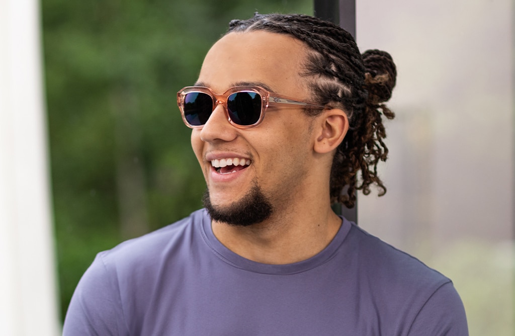 Perri Kiely smiling outdoors in bright daylight, wearing a blue t-shirt and Transitions® GEN S™ lenses, with his hair tied back.