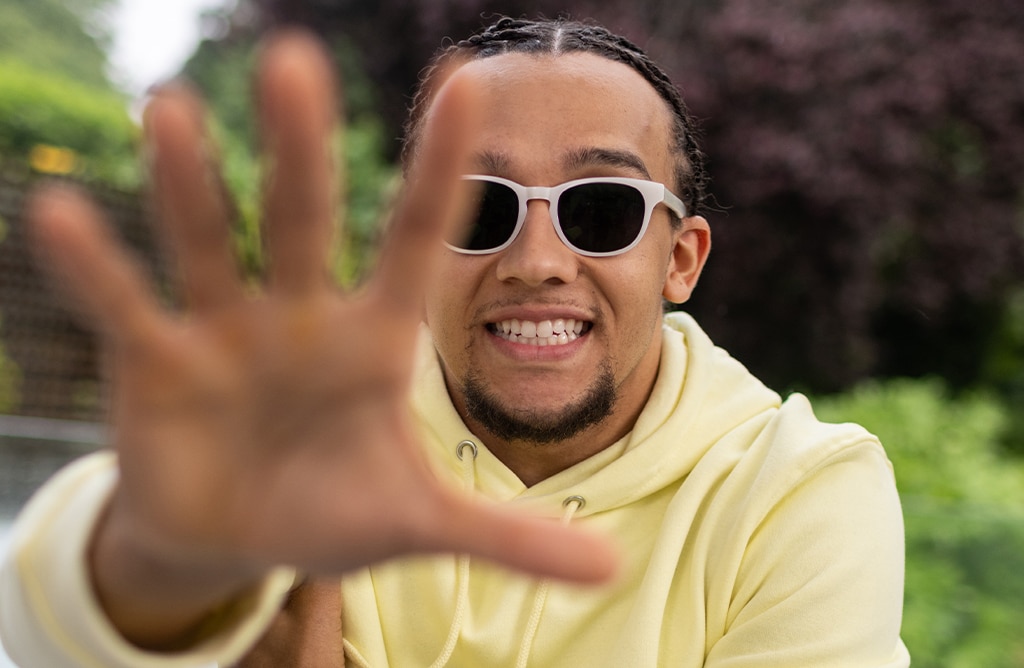 Perri Kiely outdoors in bright light, hair tied back, wearing a pastel yellow jumper and sapphire Transitions® lenses, with his hand partially covering the camera.