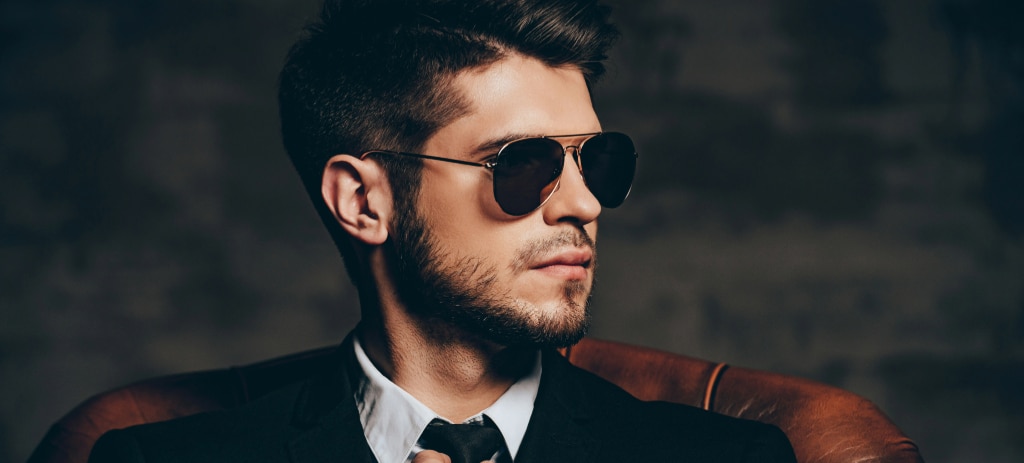 A man with a quiff sits in a dark setting on a leather chair, gazing to his left. He wears a black suit and tie paired with gold Ray-Ban Aviator sunglasses.