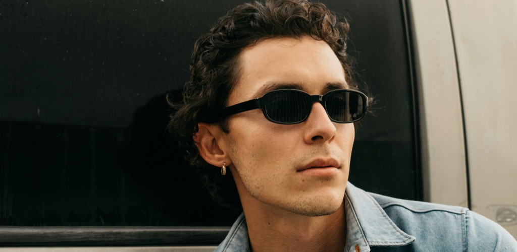 Close-up of a man with short curly dark hair wearing narrow black 90s-style sunglasses and a small hoop earring, leaning against a vehicle window in a light denim jacket, looking off to the side.