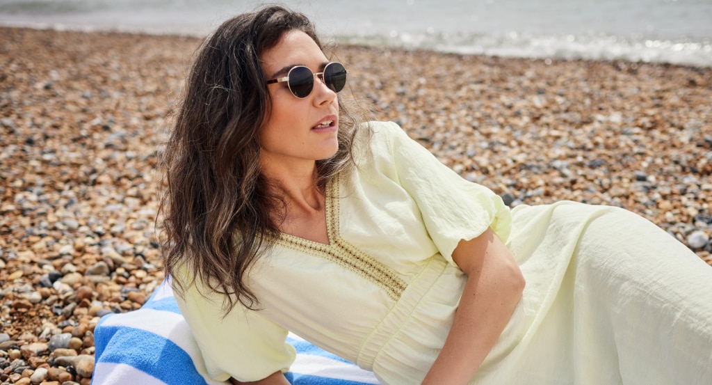 A young brunette woman wearing small round metal sunglasses and a pale yellow summer dress reclines on a striped blue and white towel on a pebble beach, gazing to the side with the sea softly out of focus in the background.