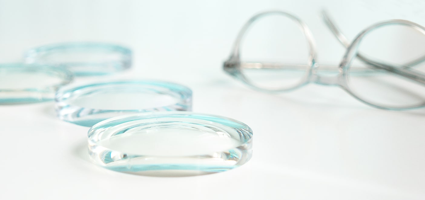 Close-up of thick eyeglass lenses placed on a surface with a pair of round transparent glasses blurred in the background.