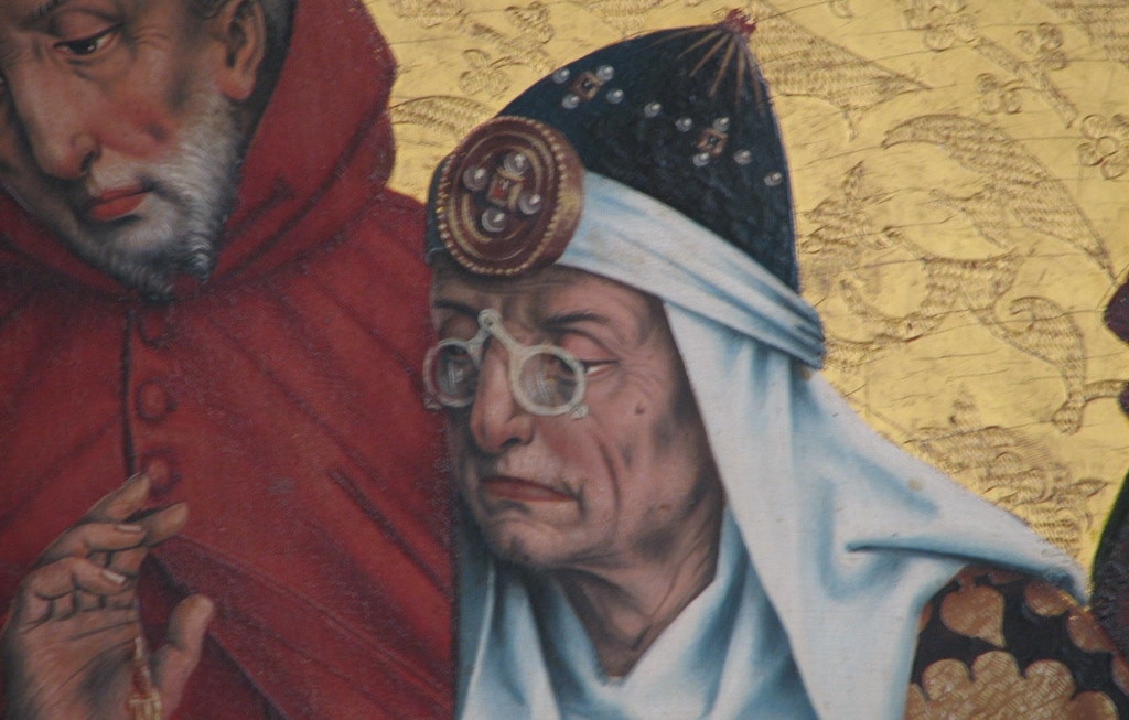 A medieval illustration showing an old man with a middle-eastern head-dress wearing scissor glasses on his nose