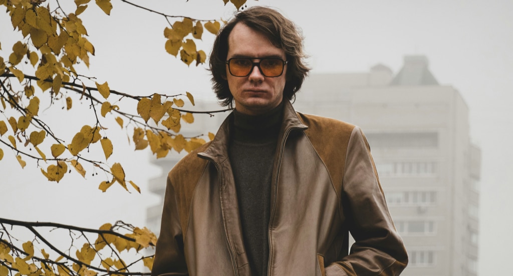 A man with medium-length brown hair wearing amber-tinted aviator sunglasses, a dark turtleneck, and a brown leather jacket stands outdoors beneath autumn leaves, with a misty urban building blurred in the background.