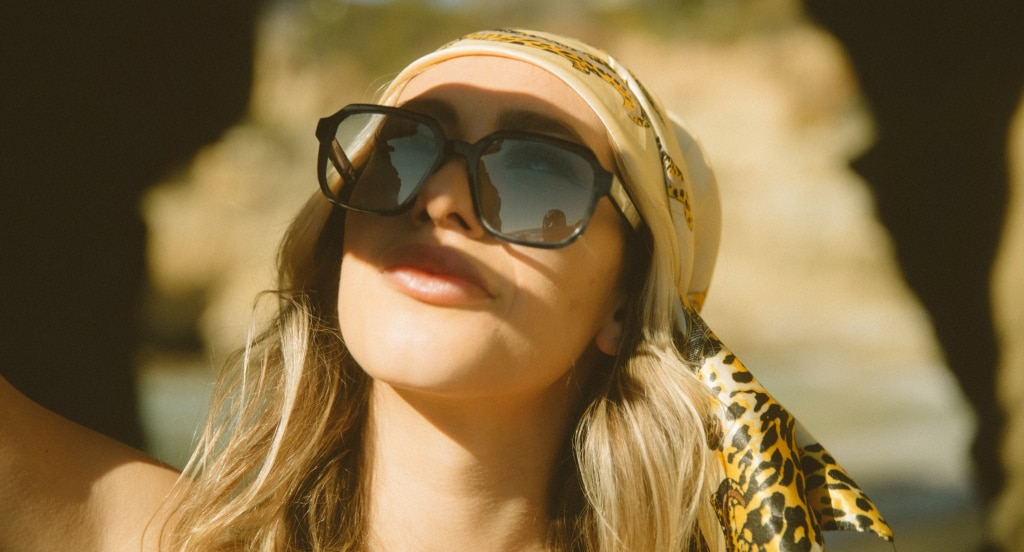 A young blonde woman wearing oversized black sunglasses and a patterned headscarf looks upward in warm sunlight, with a rocky coastal arch and water softly blurred in the background.