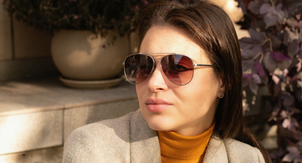 A young woman with long brown hair wearing aviator-style sunglasses with tinted lenses, an orange turtleneck, and a neutral-toned coat looks to the side in soft sunlight, with potted plants and foliage blurred in the background.