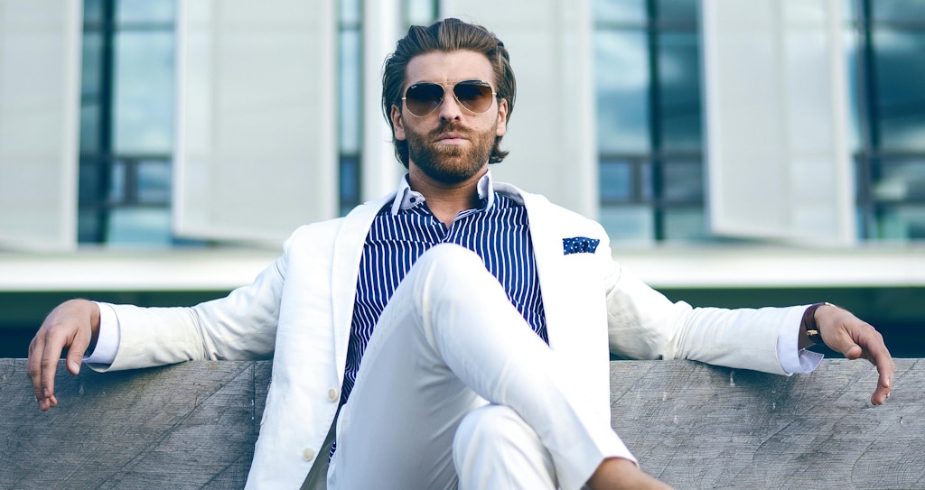 A man with slicked back hair wearing a white suit and aviator sunglasses, sitting on a bench outdoors with his legs crossed