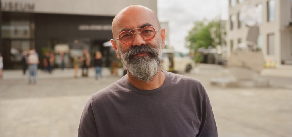 Bald man with a grey beard wearing round red glasses with metal accents and a blue long-sleeve top, standing outdoors in an urban area with buildings and people blurred in the background.
