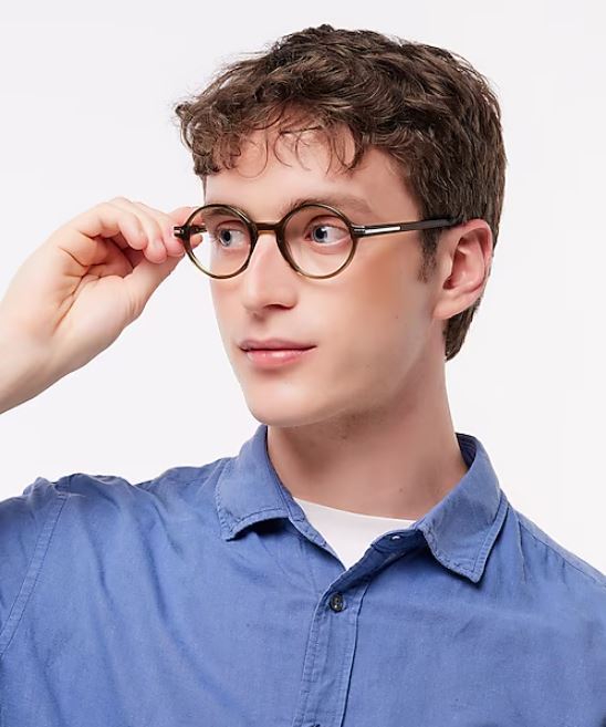 Man with short curly brown hair wearing round olive-green glasses, dressed in a blue button-up shirt over a white t-shirt, looking to the side while adjusting his frames on a neutral background.