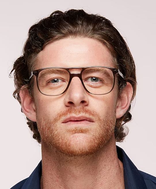 Close-up of a man with wavy brown hair and light facial hair wearing large rectangular grey glasses with a unique bridge design, looking straight at the camera against a neutral background.