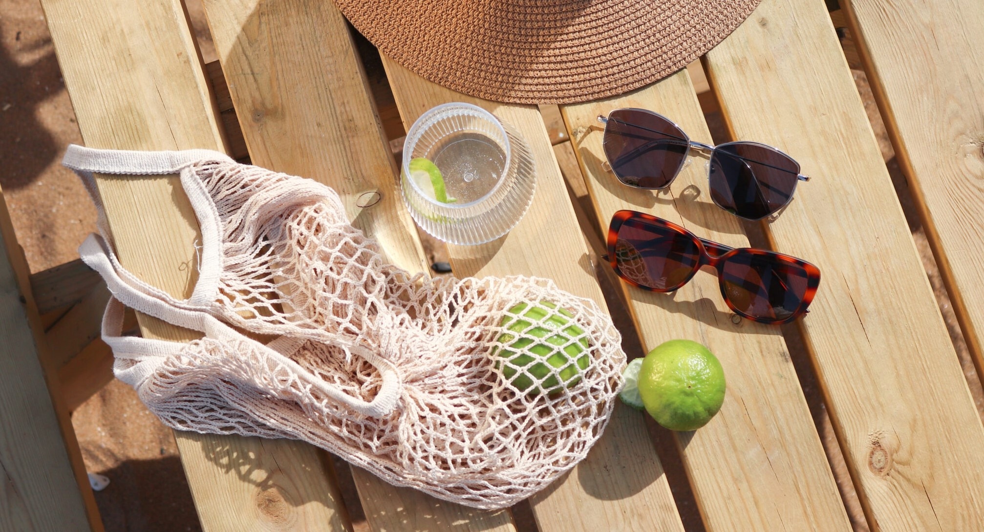 The brim of a hat, netted-bag, limes, glass of water and two pairs of sunglasses on a bench in a bright beach location