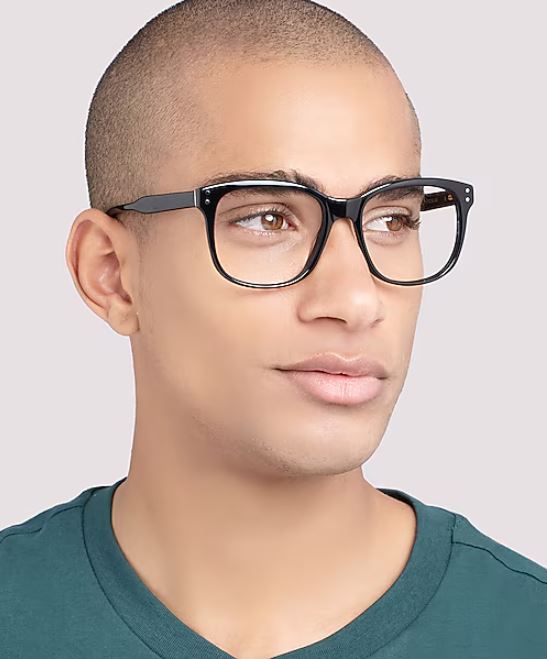 Close-up of a man with a shaved head wearing thick black square glasses and a teal crew-neck shirt, looking slightly to the side against a neutral background.