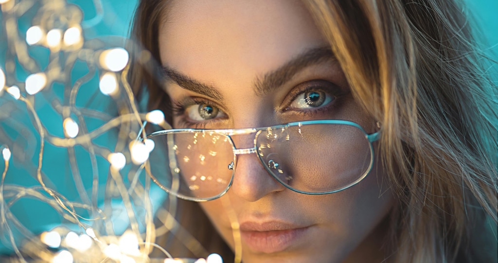 A woman with brown eyes peeking over the top of her metal glasses at a string of fairy lights