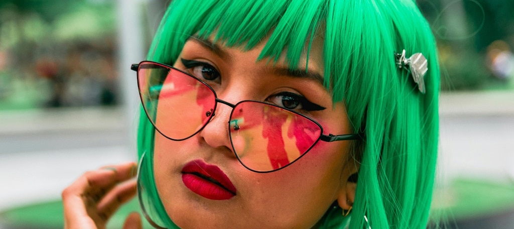 A young woman with green hair wearing thin black cat eye glasses with almost triangular lenses with a pink tint