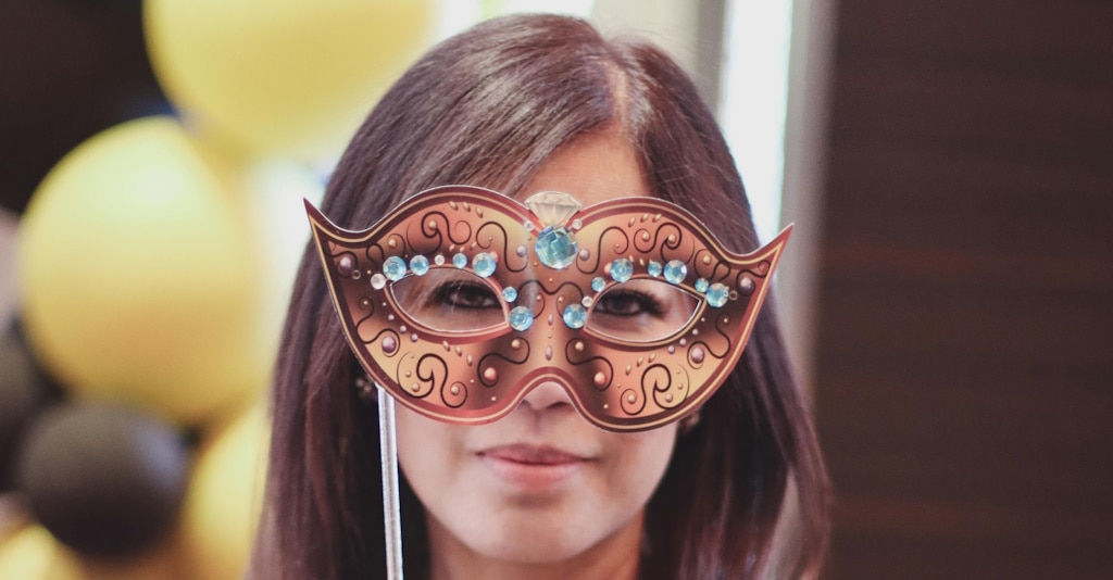 A young woman with straight brown hair holds an imitation of a golden, gem-encrusted Venetian-style mask up to her face