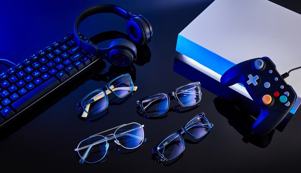 Four pairs of glasses with their lenses reflecting blue light lying on a shiny dark table, next to a keyboard, a pair of wireless headphones and a gaming controller