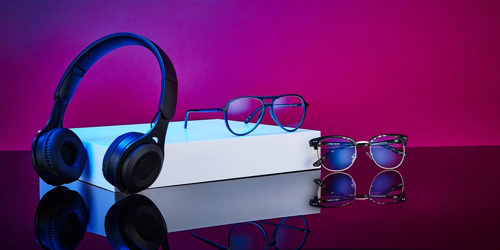 Two pairs of glasses and a pair of wireless headphones lying on a smooth, reflective surface with a bright purple backdrop