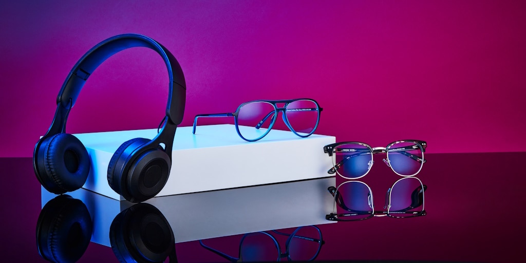 A pair of wireless black overhead headphones and two pairs of modern eyeglasses displayed on a reflective surface, with a white block stand and a purple–blue gradient background.