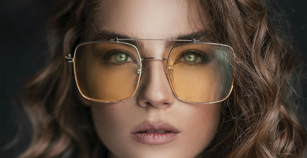 A woman with green eyes and wavy brown hair, staring intently at the viewer through a pair of square aviator glasses with lightly tinted lenses