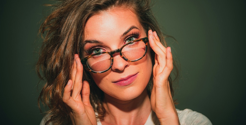 A woman with wavy hair raising her eyebrows as she peeks over the top of her tortoiseshell glasses and they cast shadows on her face