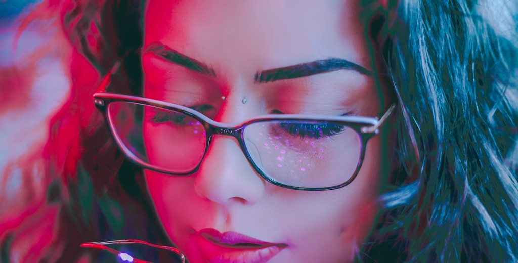 Close-up of a woman with thick eyelashes and wearing thin square tortoiseshell glasses while bathed in pink light