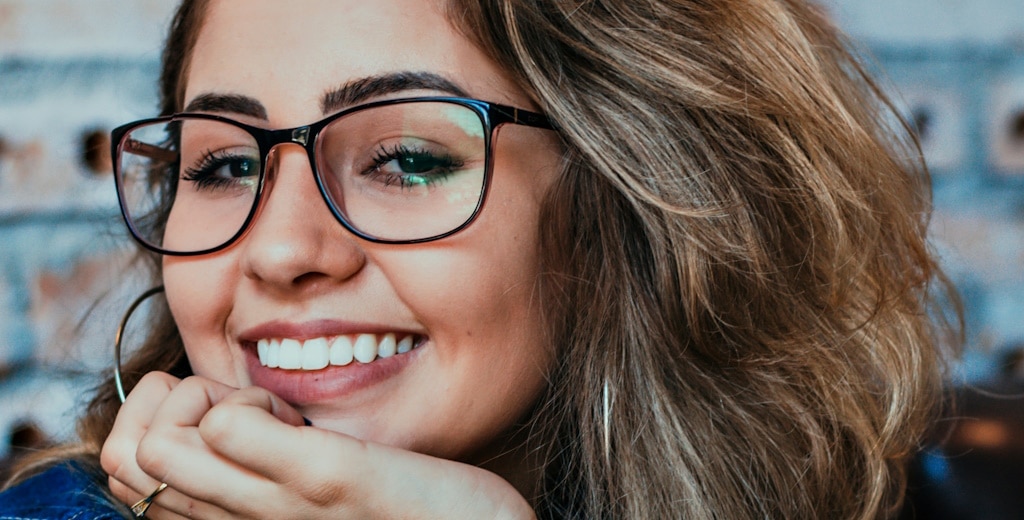 A smailing woman wearing square black glasses and makeup that highlights her eyes and brows