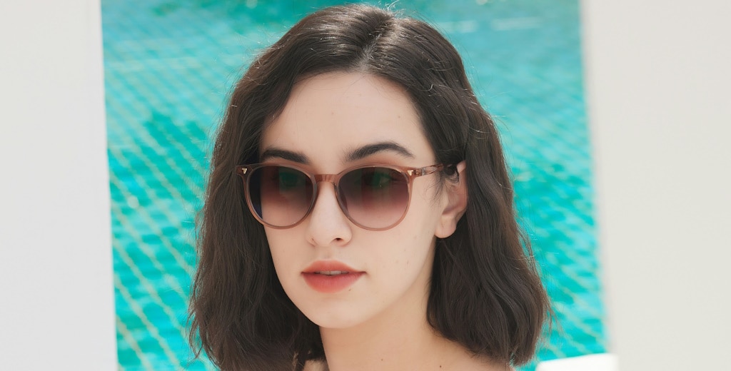 A woman with straight brown hair wearing light pink sunglasses and light red lipstick by a pool