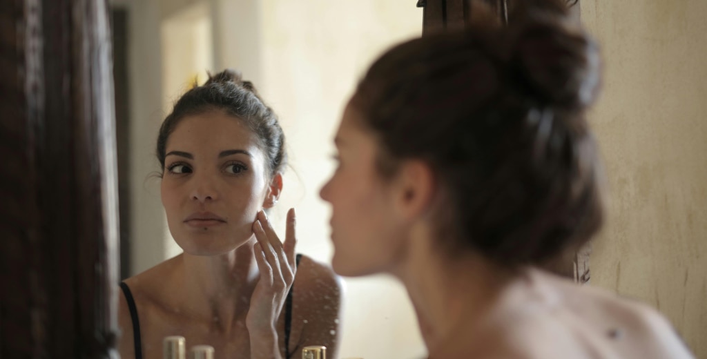 A lighly dressed woman looking into a mirror while applying skincare to her cheek