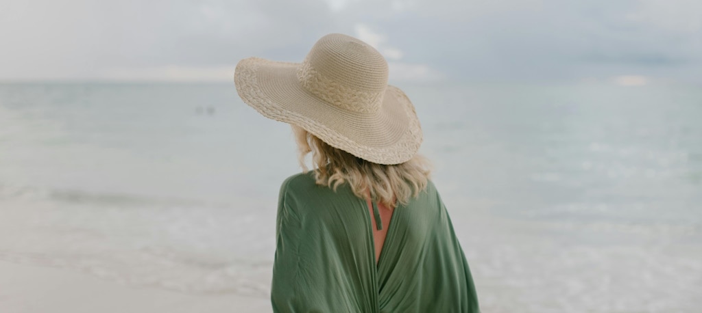 View from behind of a blonde woman with a sun hat and wearing a green linen shirt walking along a beach