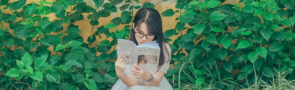 Viatnamese young woman wearing black glasses reading a book while sitting outside among plants in front of a house