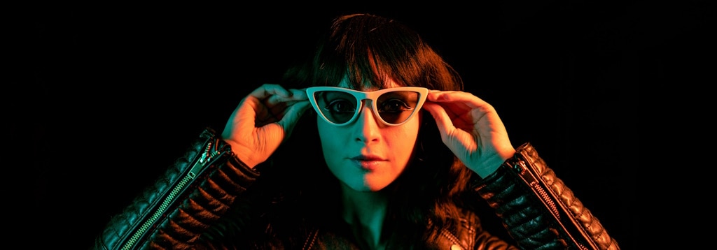 A woman in a black leather jacket wearing a pair of white cat-eye sunglasses while bathed in a reddish glow